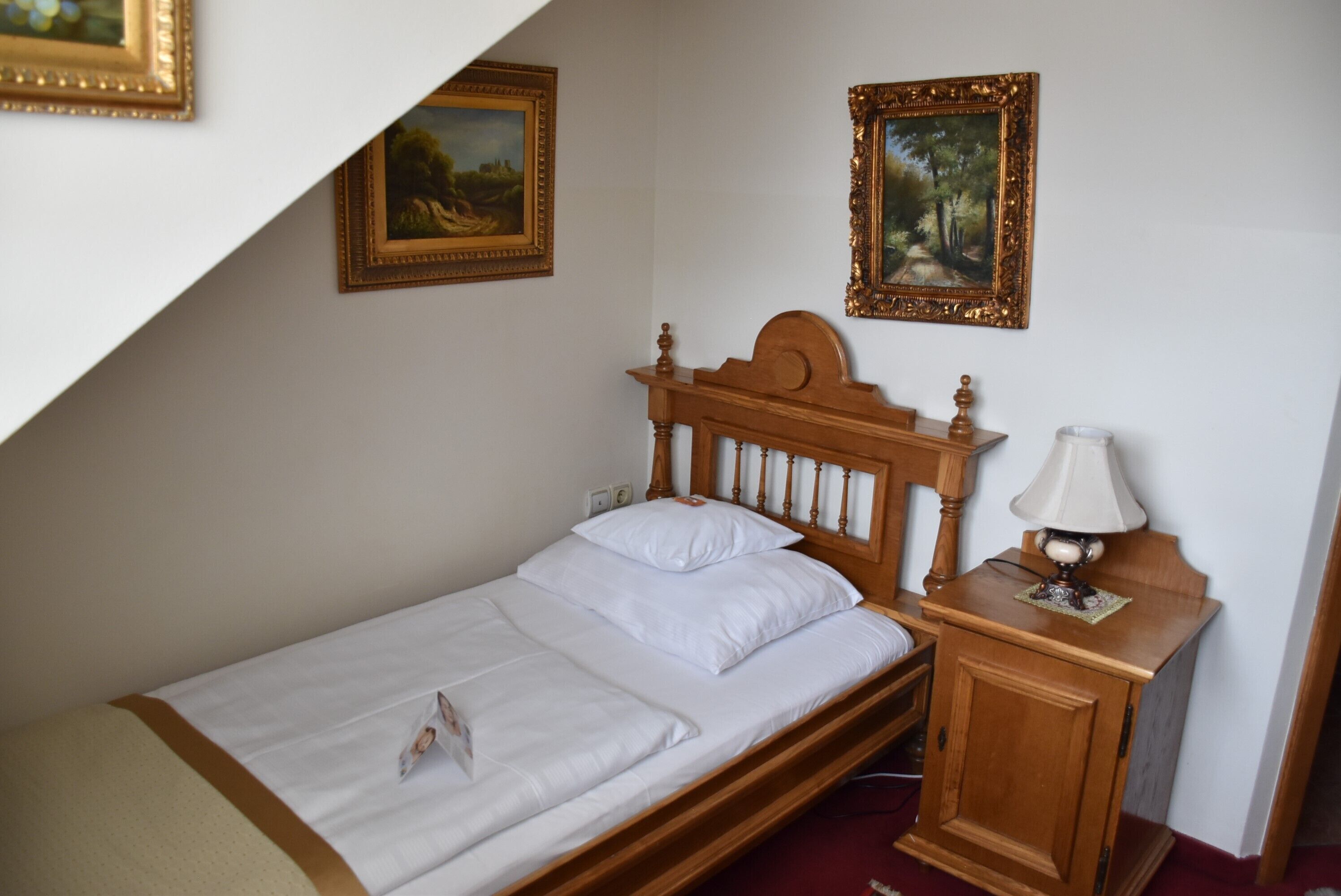 A single bed with a wooden headboard, a bedside table with a lamp, and two framed paintings on the wall.