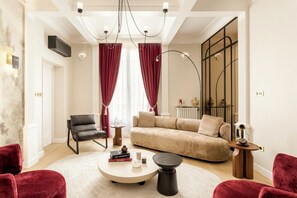 Una sala de estar moderna con un sofá color beige, una silla negra y sillones de terciopelo rojo. Hay una mesa de centro redonda con libros y velas.