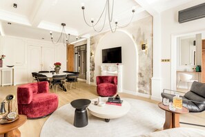 A spacious living room with a round coffee table, red velvet armchairs, a black sofa, and a dining area with a white table and black chairs.
