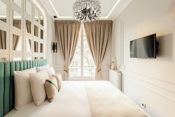 A modern bedroom with a green upholstered headboard, a large bed with white linens, beige curtains, and a mounted TV on the wall.