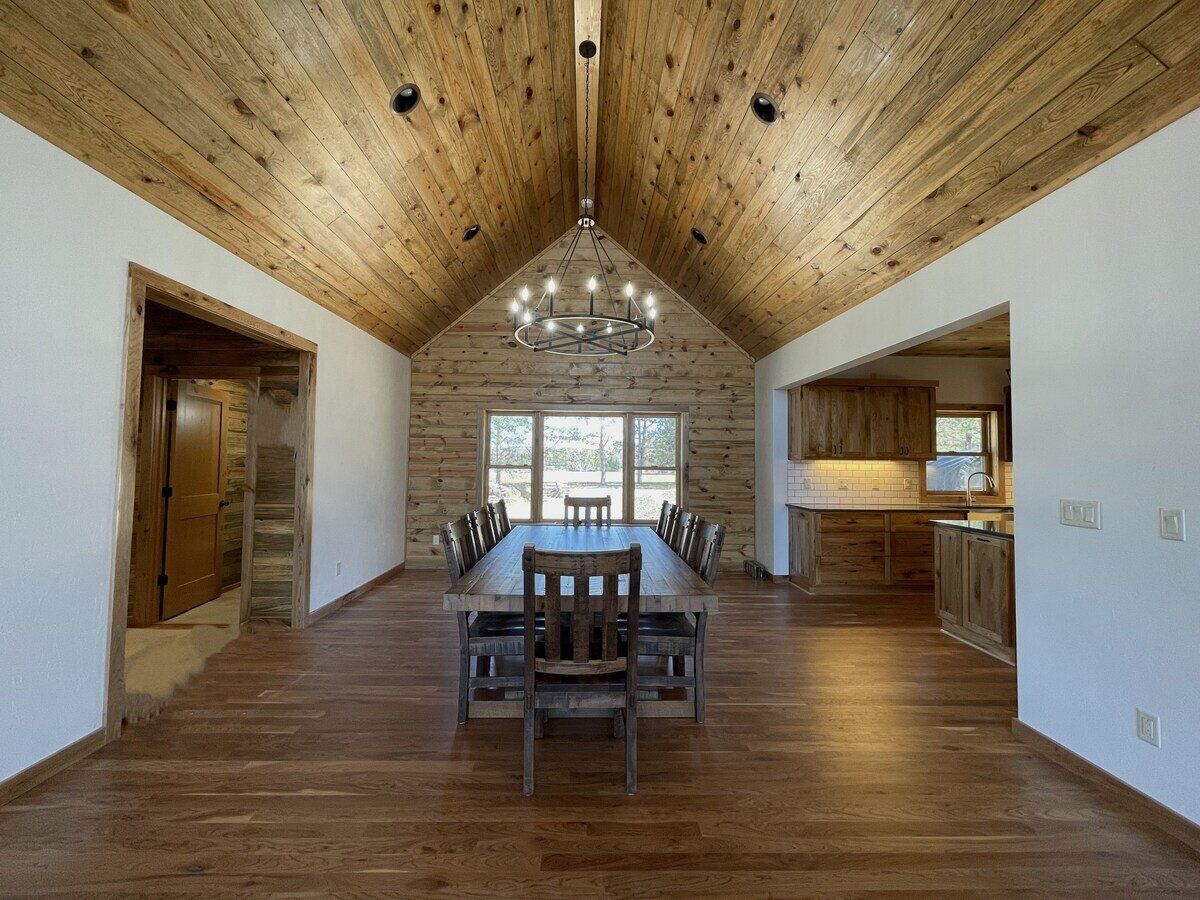Une salle à manger spacieuse, avec une longue table et des chaises en bois, un haut plafond également en bois, et une vue dégagée grâce à de grandes fenêtres.