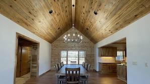 A spacious dining area with a long wooden table and chairs, a high wooden ceiling, and a view through large windows.