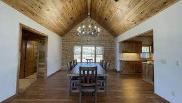 Une salle à manger spacieuse, avec une longue table et des chaises en bois, un haut plafond également en bois, et une vue dégagée grâce à de grandes fenêtres.