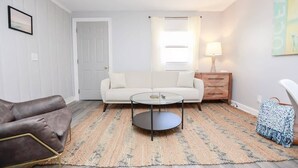 A living room with a white sofa, a brown leather chair, a glass coffee table, a wooden side table with a lamp, and a patterned rug. - Sojourns Spit 2 BR  Beach Shack 1278WOV#2 (Norfolk)