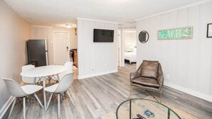 A modern living room with a white dining table and chairs, a brown leather sofa, a flat-screen TV mounted on the wall, and a 'BEACH' sign. - Sojourns Spit 2 BR  Beach Shack 1278WOV#2 (Norfolk)