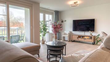 A modern living room with a sofa, a coffee table, and a TV mounted on the wall. There is a sliding glass door leading to an outdoor area with chairs.
