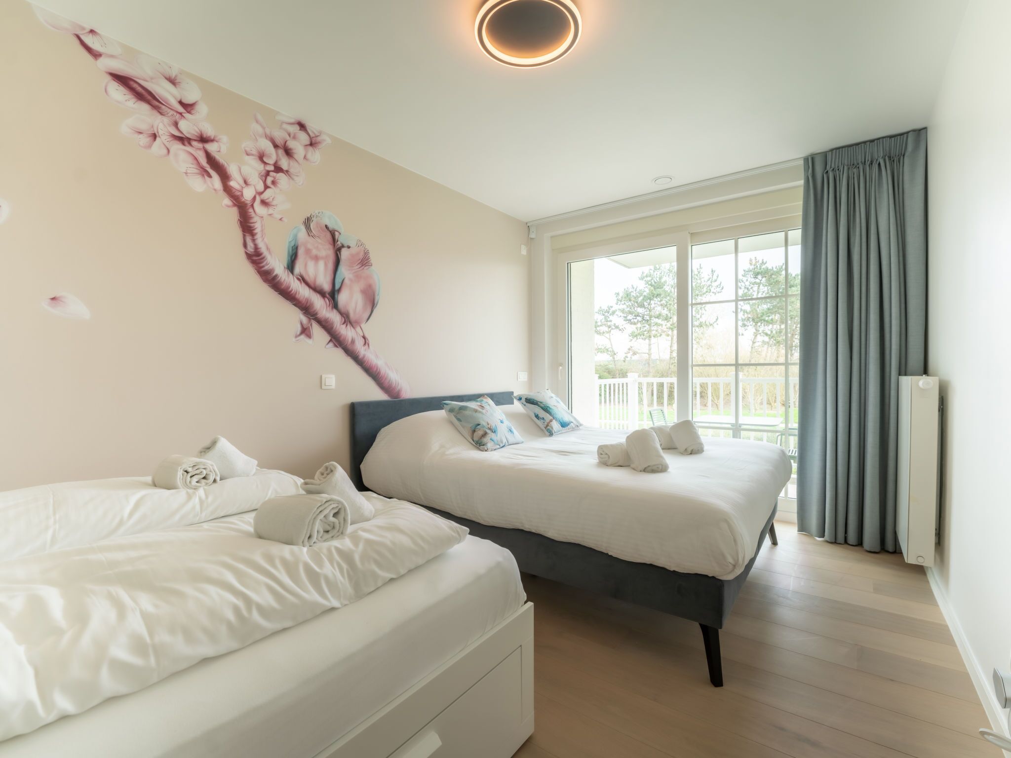 A hotel room with two beds, a large window with curtains, a wall mural, and a ceiling light.