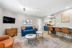 A modern living room with a sofa, coffee table, TV, and a kitchen area with bar stools.
