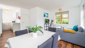 Uma sala de estar moderna e bem iluminada, com um sofá cinza, uma mesa de jantar com cadeiras, uma TV e uma janela com vista para o verde.