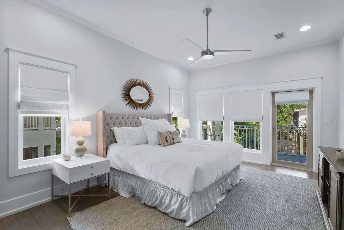 A bedroom with a large bed, a ceiling fan, a window with blinds, and a view of the outdoors.
