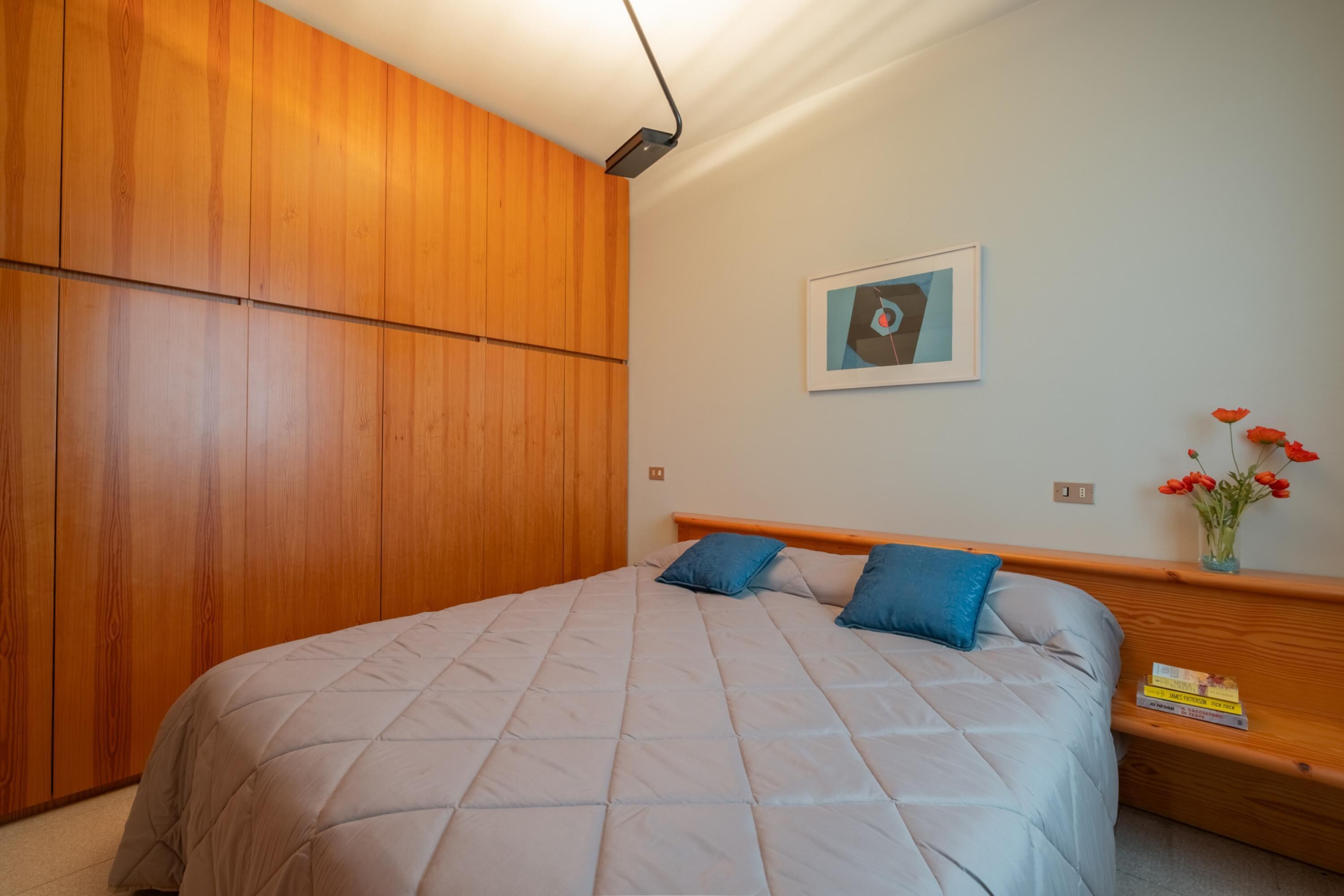 A bedroom with a bed, wooden wardrobe, a framed picture on the wall, and a bedside table with books and a vase of flowers.