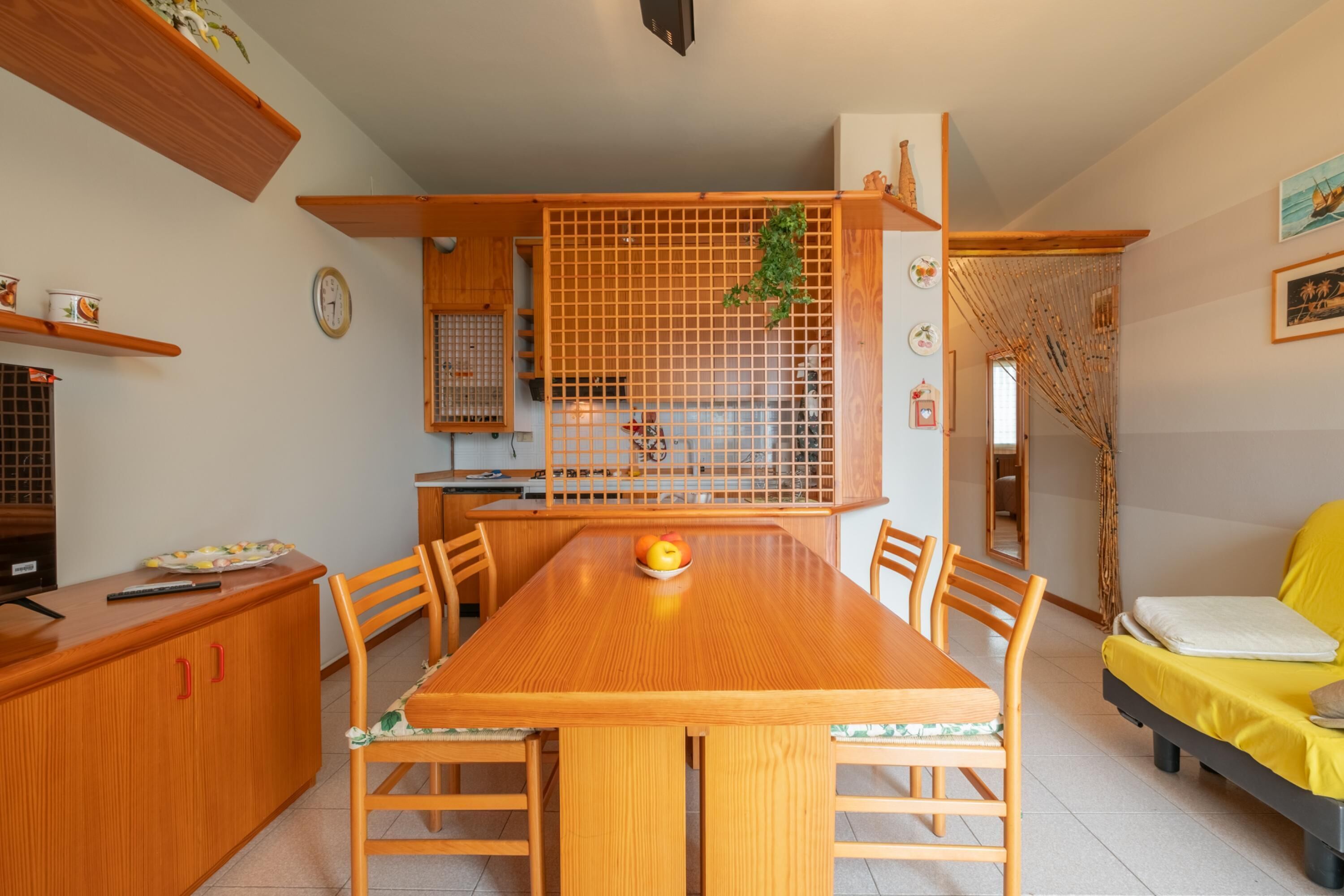 Un comedor con una mesa de madera, sillas y un frutero. Hay una zona de cocina con armarios de madera y una ventana.