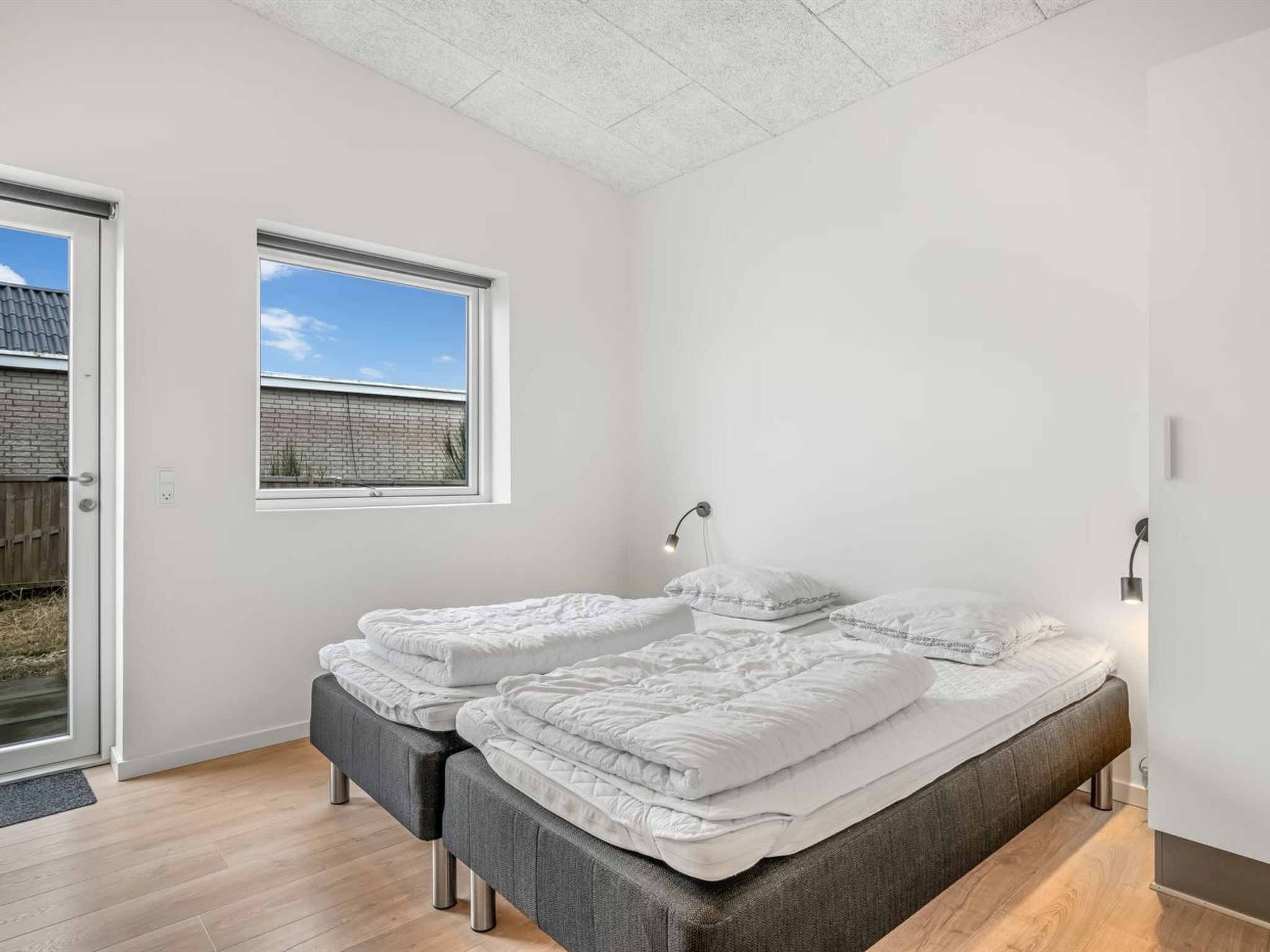 A bedroom with two beds, a window, a door, and a wall-mounted lamp.