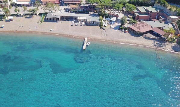 An aerial view of a coastal area with clear turquoise waters, a pier, and buildings with a mix of architectural styles.
