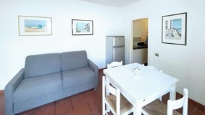 A small living room with a grey sofa, a white table with four chairs, and a refrigerator. There are framed pictures on the walls. - Sant'Anna Volterraio - Maestra 56 - Sant'Anna del Volterraio - Maestra 56 (Rio dell'Elba)