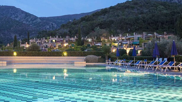 A swimming pool with checkered tiles, lounge chairs, and umbrellas, surrounded by a mountainous landscape with buildings in the background. - Sant'Anna Volterraio - Maestra 56 - Sant'Anna del Volterraio - Maestra 56 (Rio dell'Elba)