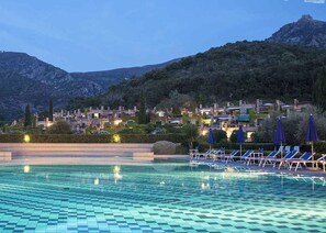A swimming pool with checkered tiles, lounge chairs, and umbrellas, surrounded by a mountainous landscape with buildings in the background.