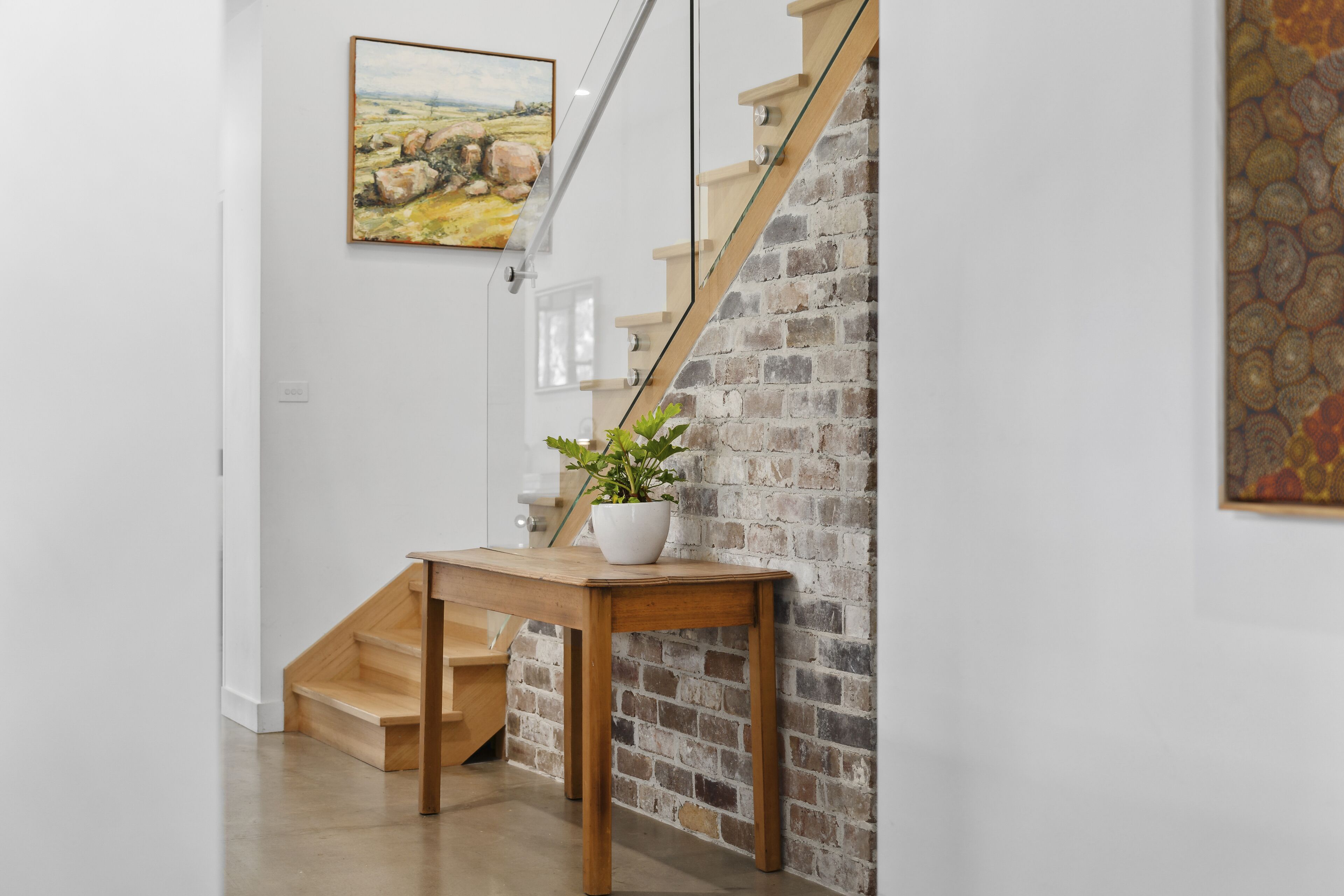 Een trap met houten treden en een bakstenen muur, een houten tafel met een potplant en een schilderij aan de muur.
