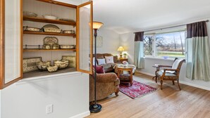 Ein Wohnzimmer mit einer Couch, Sesseln, einem Couchtisch, einer Stehlampe, einem Bücherregal und einem Fenster mit Vorhängen.