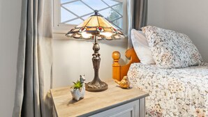 Ein Bett mit floraler Bettwäsche, eine Nachttischlampe, eine kleine Pflanze und ein dekorativer Gegenstand auf einem Holznachttisch.