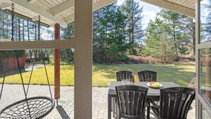 Une terrasse couverte avec une table et des chaises, une balançoire suspendue et une vue sur un espace verdoyant avec des arbres.