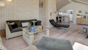 Un vaste espace de vie comprenant un canapé, une table basse en verre et un fauteuil en cuir. On aperçoit une cuisine au fond.