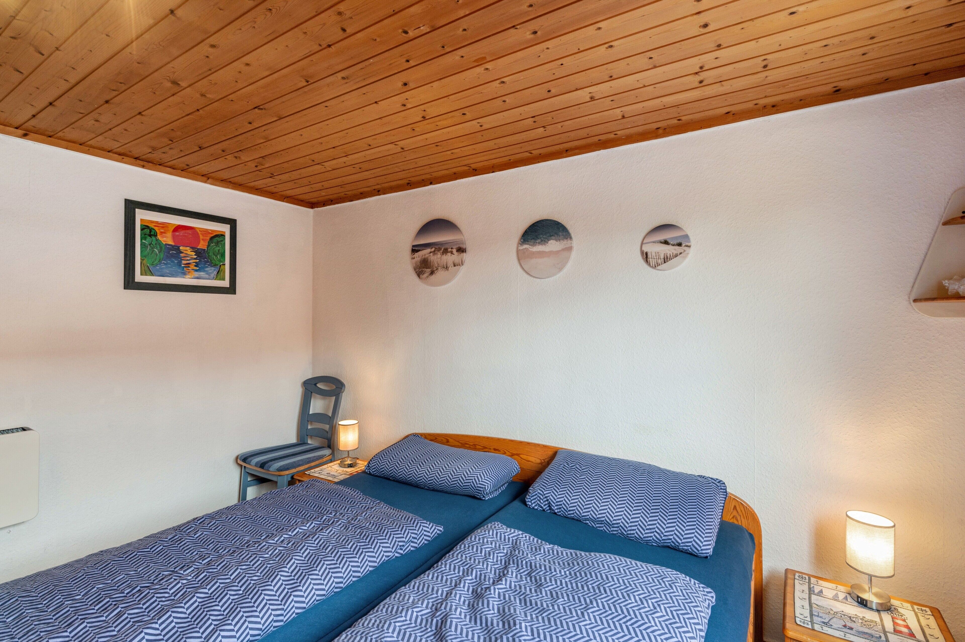 Een slaapkamer met twee bedden, een houten plafond, een wandradiator, een stoel en een ingelijst kunstwerk.