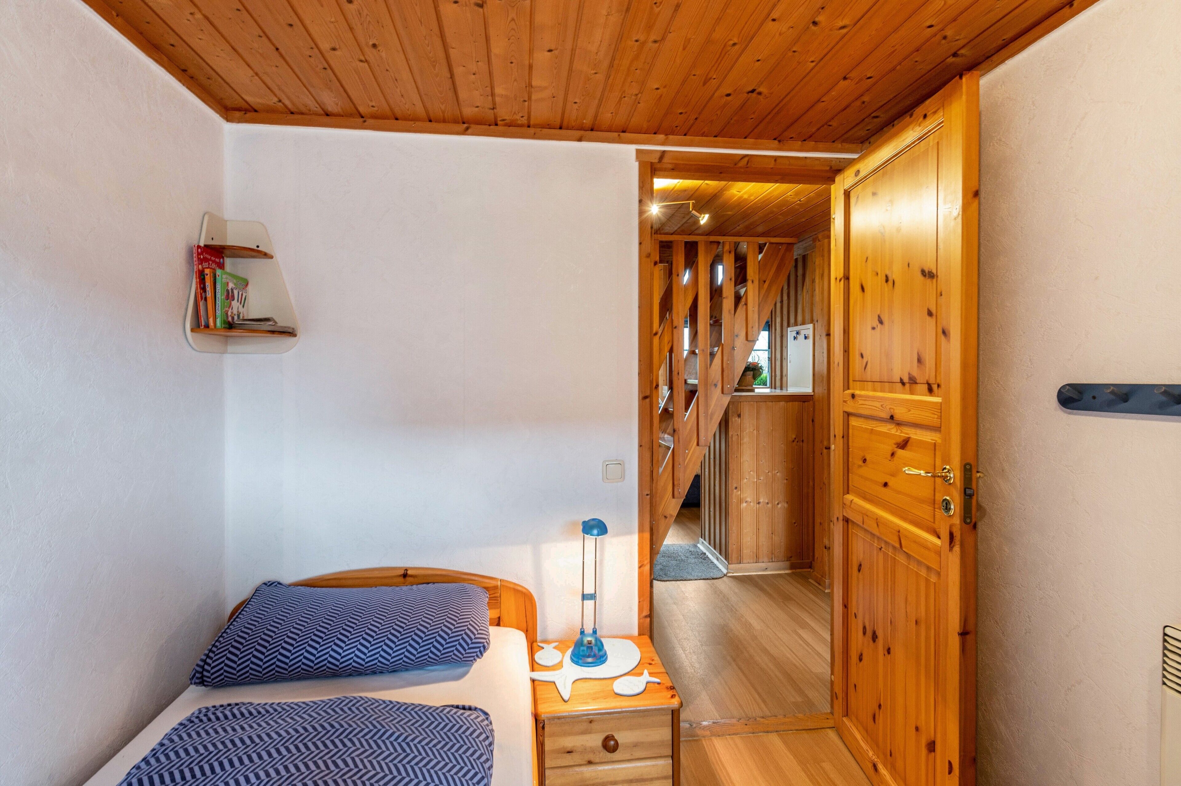 Quarto com teto de madeira, cama com roupa de cama estampada, mesa de cabeceira com luminária e guarda-roupa de madeira.