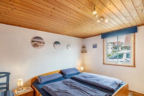 A bedroom with a wooden ceiling, a bed with patterned bedding, a nightstand with a lamp, and a window with a view of buildings and a car.