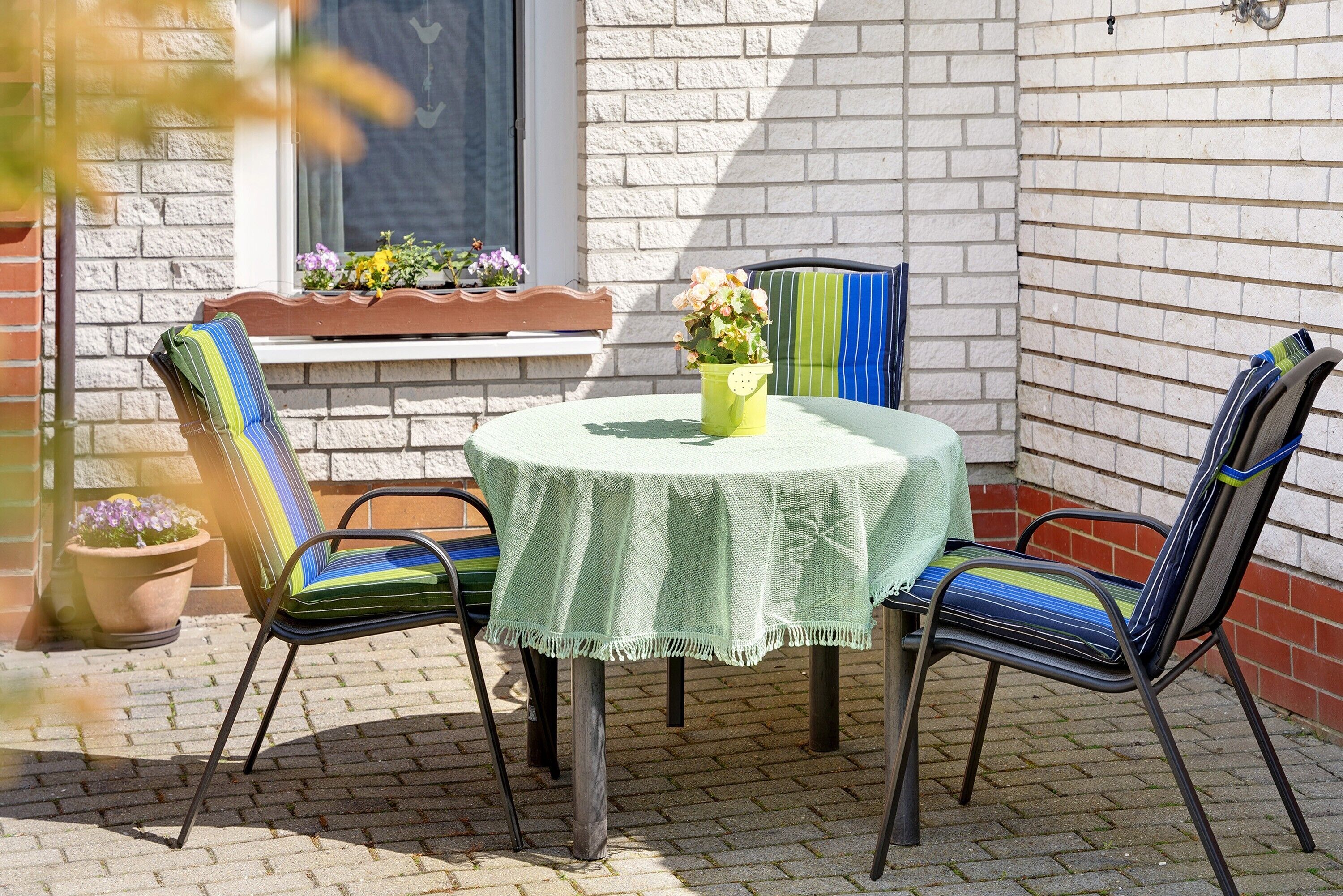 A patio with a round table, two striped chairs, a potted plant, and a window with a view.
