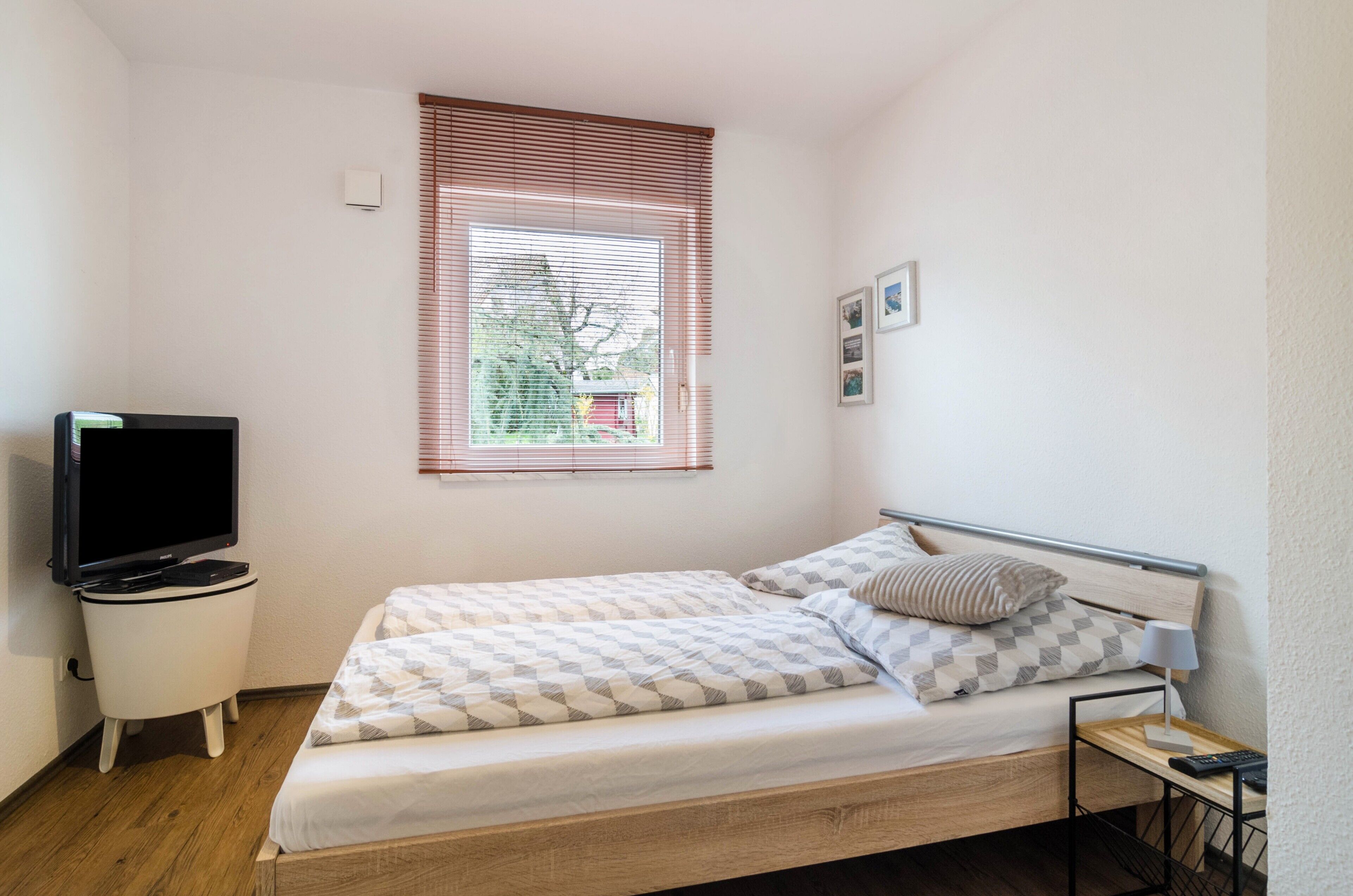 Ein kleines, ordentlich eingerichtetes Schlafzimmer mit einem Einzelbett, einem Fernseher, einem Nachttisch und einem Fenster mit Jalousien.