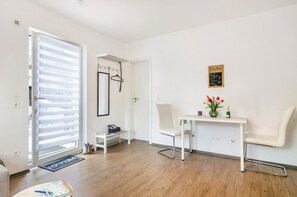 Une pièce moderne et bien éclairée, avec un plancher en bois, une chaise blanche, une petite table ornée d’un vase de fleurs et une porte donnant sur l’extérieur.