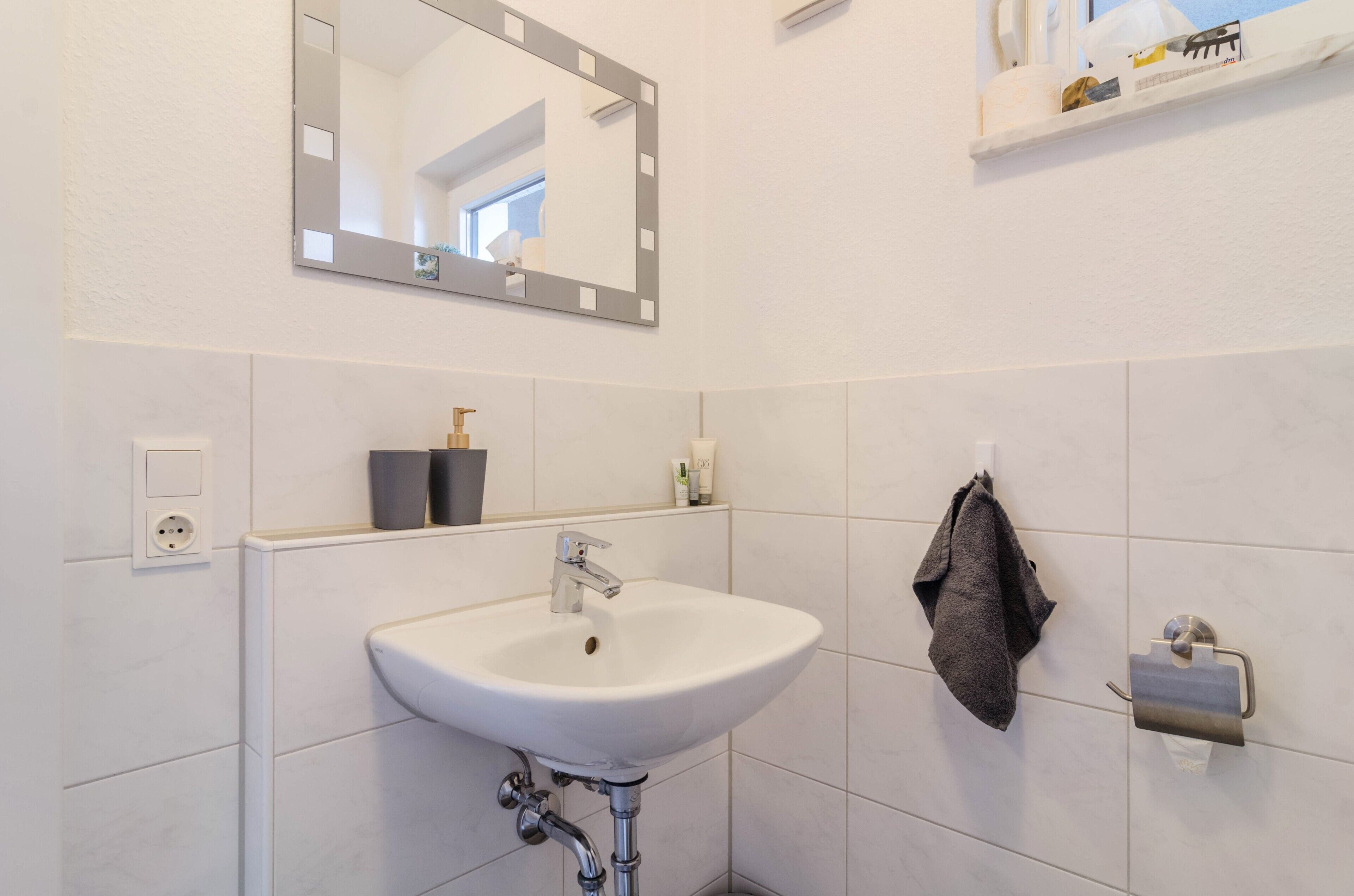Un bany amb un lavabo blanc, un mirall, una barra de tovalloles i un suport de paper higiènic.