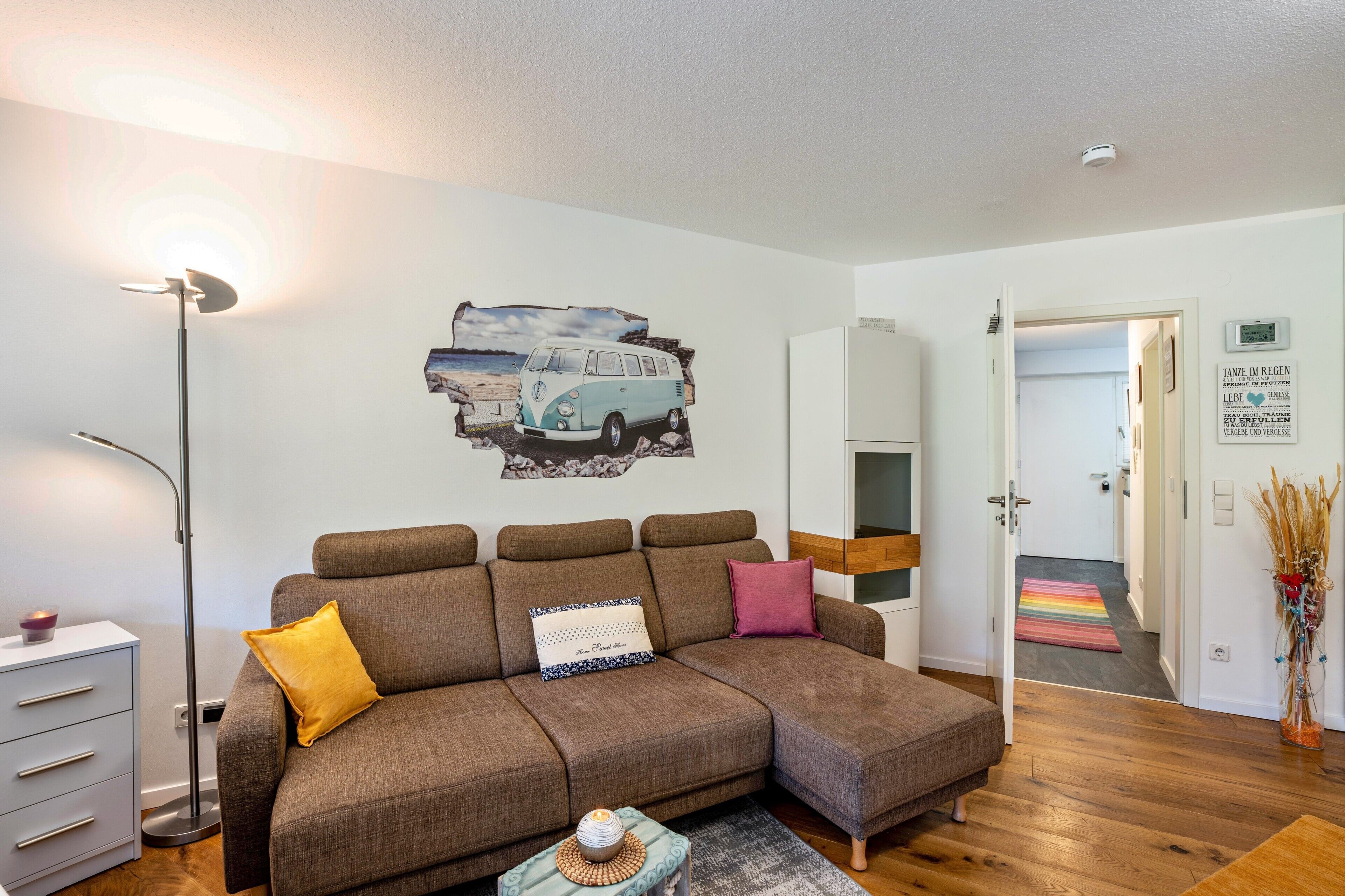 Un salon moderne avec un canapé, une lampe, une décoration murale représentant un fourgon, et une porte donnant sur une autre pièce.