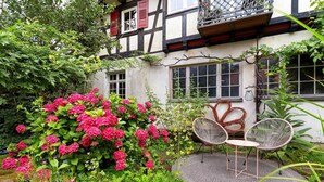 Un jardín con una mesa y sillas, una casa con balcón y una ventana con contraventanas.