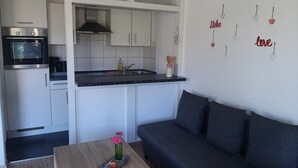 Une cuisine compacte avec des meubles blancs, un four encastré et une machine à café. Une petite table à manger avec un vase de fleurs se trouve au premier plan.