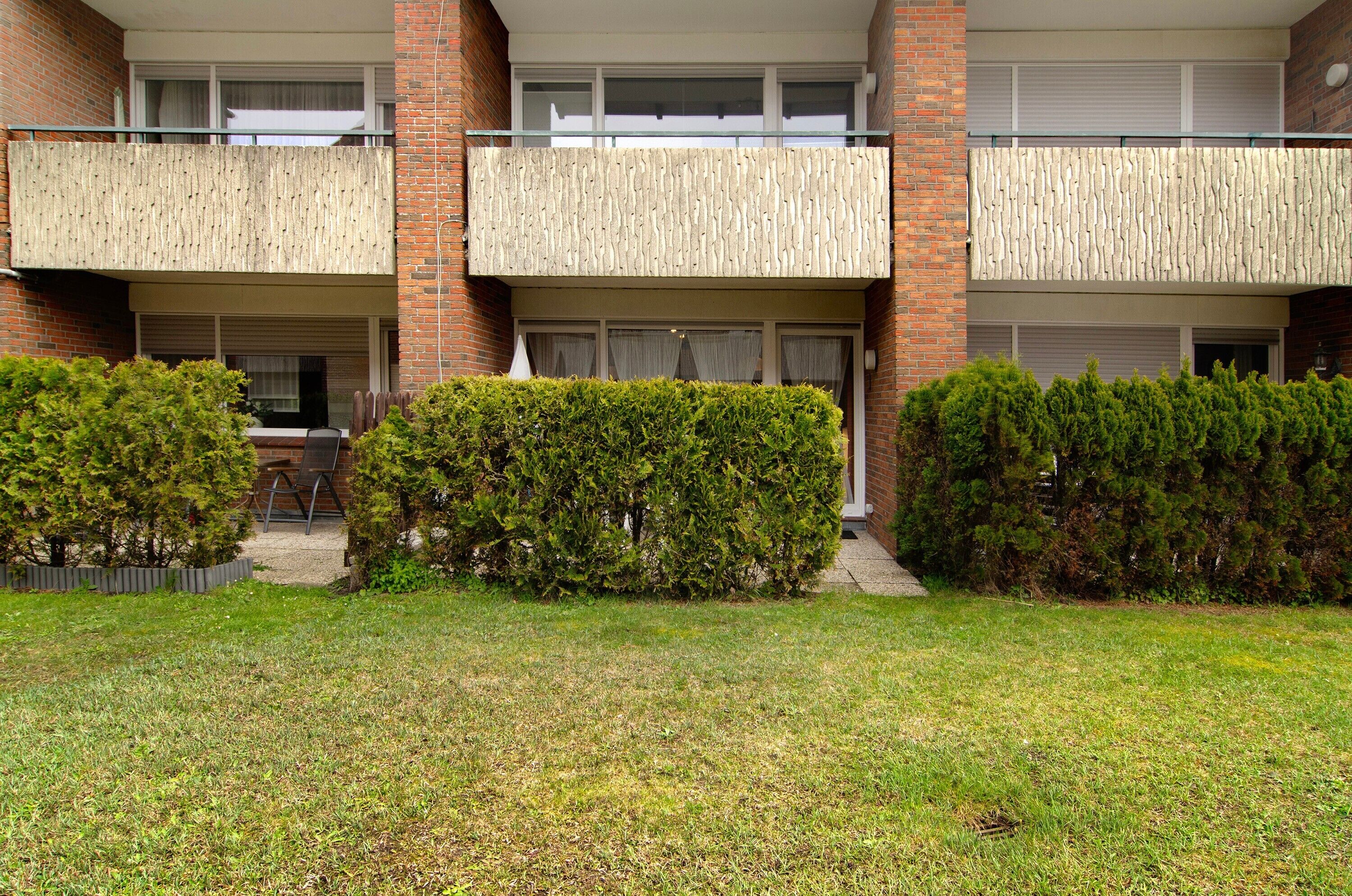 Ein zweistöckiges Gebäude mit Flachdach, einem Balkon im oberen Stockwerk und einer kleinen Gartenanlage mit geschnittenen Büschen.