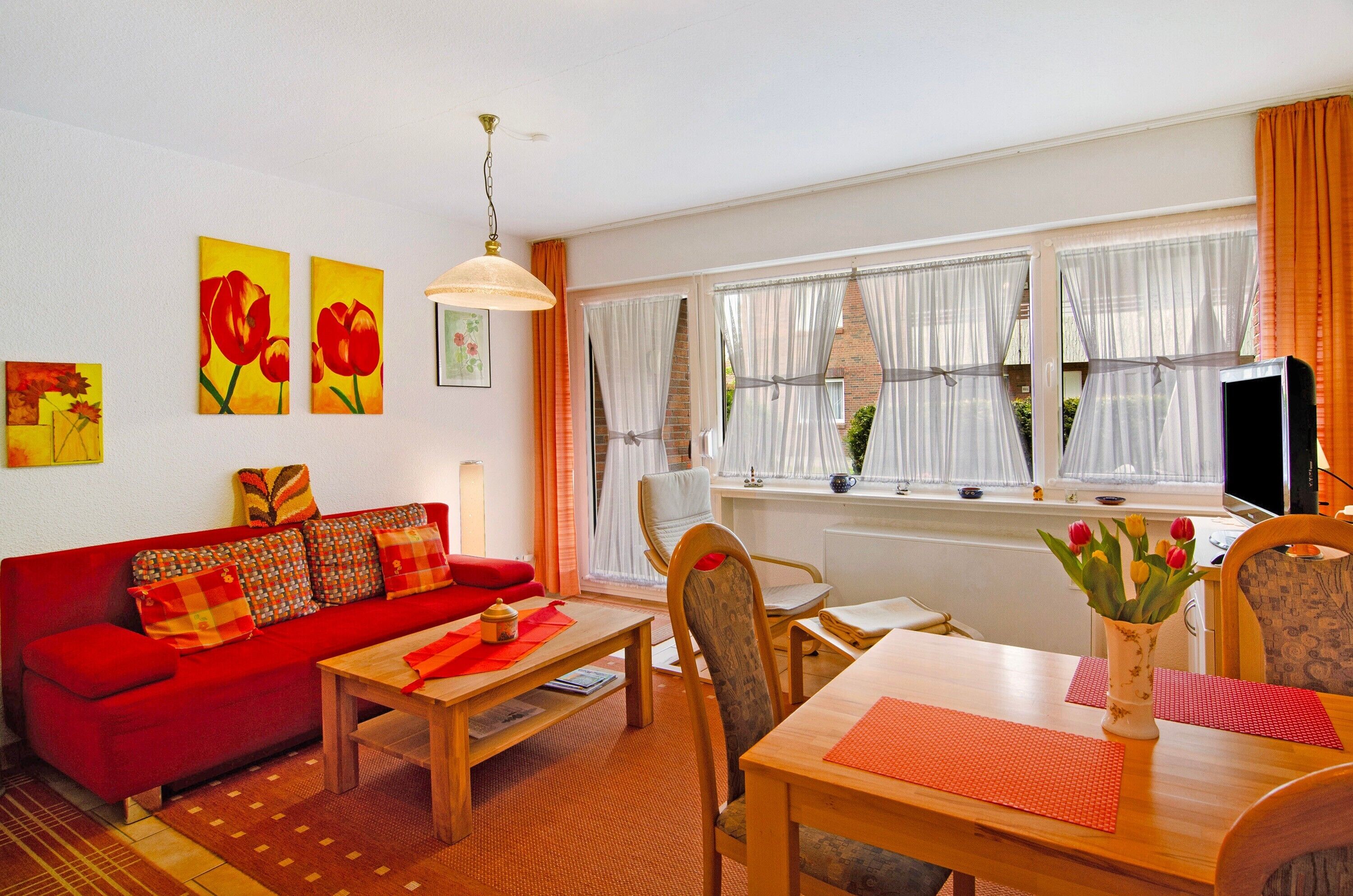 Ein Wohnzimmer mit einem roten Sofa, einem Holztisch und einem Esstisch mit Stühlen. An der Wand hängen zwei Gemälde, auf dem Esstisch steht eine Vase mit Blumen, und ein Fenster mit Vorhängen.