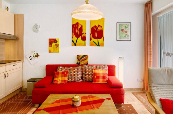 A red sofa with patterned cushions, a wooden table with a decorative item, a wall clock, and a window with curtains.