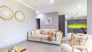 Una sala moderna con un sofá beige, una mesa de centro de madera y una mesita auxiliar con una planta. Se ve una cocina con encimera verde al fondo.