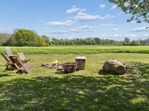 A grassy field with a fire pit, two wooden chairs, a trash bin, and a large rock.