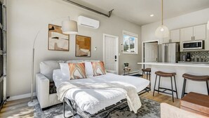 A modern bedroom with a bed, bedside tables, a desk, a chair, and a kitchen area with a refrigerator, microwave, and stools.