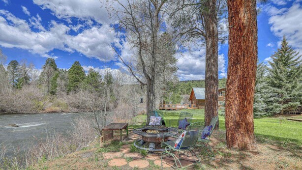 A rustic outdoor seating area with a fire pit, metal chairs, and a table, surrounded by trees and a view of a house in the background. - Riverfront Fire Pit, Gas Stove, Near Skiing, Hot Springs, & Hiking Adventures! (Pagosa Springs)