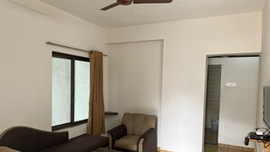 A living room with a sofa, armchair, and coffee table. There is a ceiling fan, a window with curtains, and a door leading to another room. - Ruturaj Inn (Alibag)