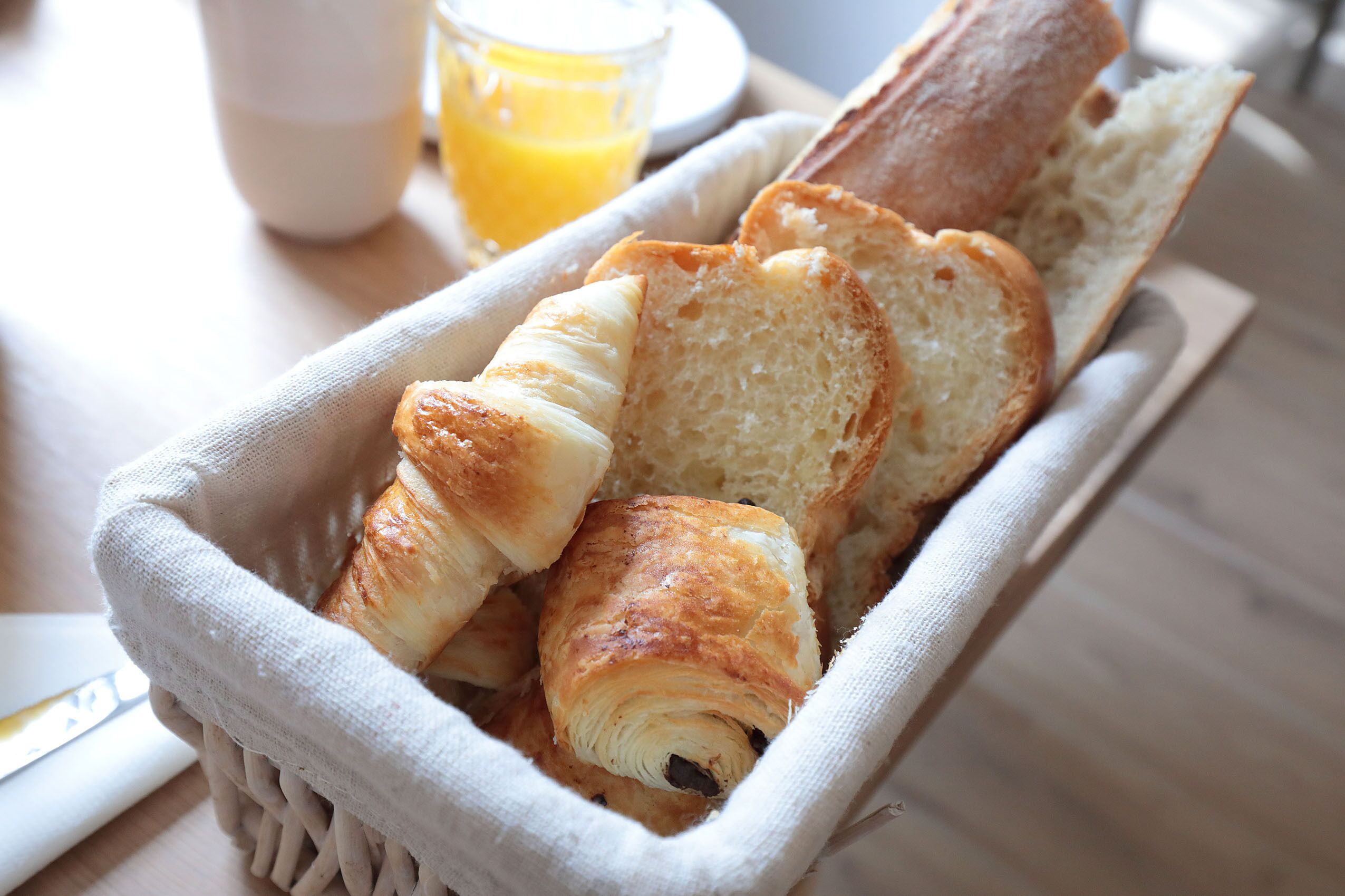 A basket containing croissants and bread slices, with a glass of orange juice and a cup in the background.