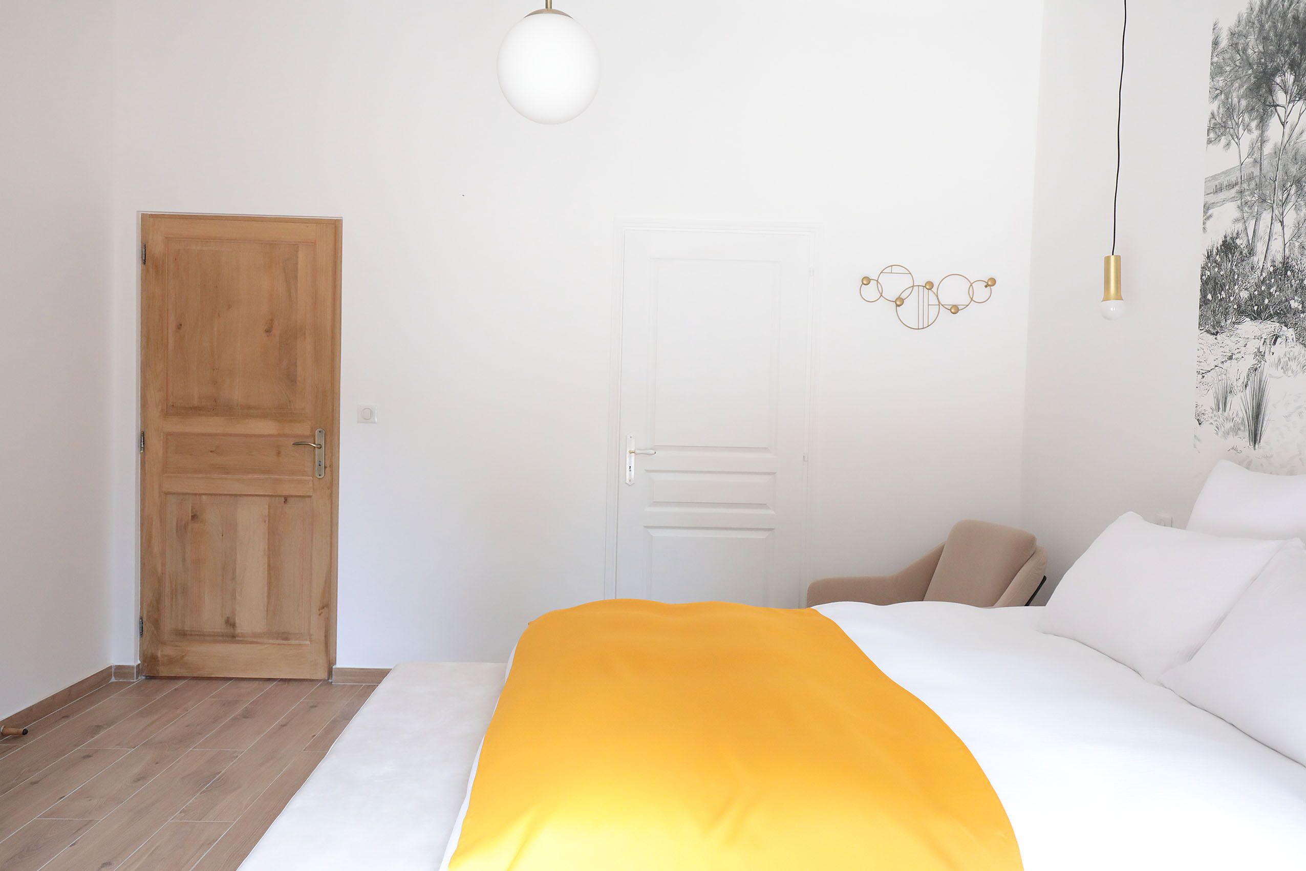A bedroom with a wooden door, a white wall, a bed with a white and orange blanket, a beige chair, and a hanging light fixture.