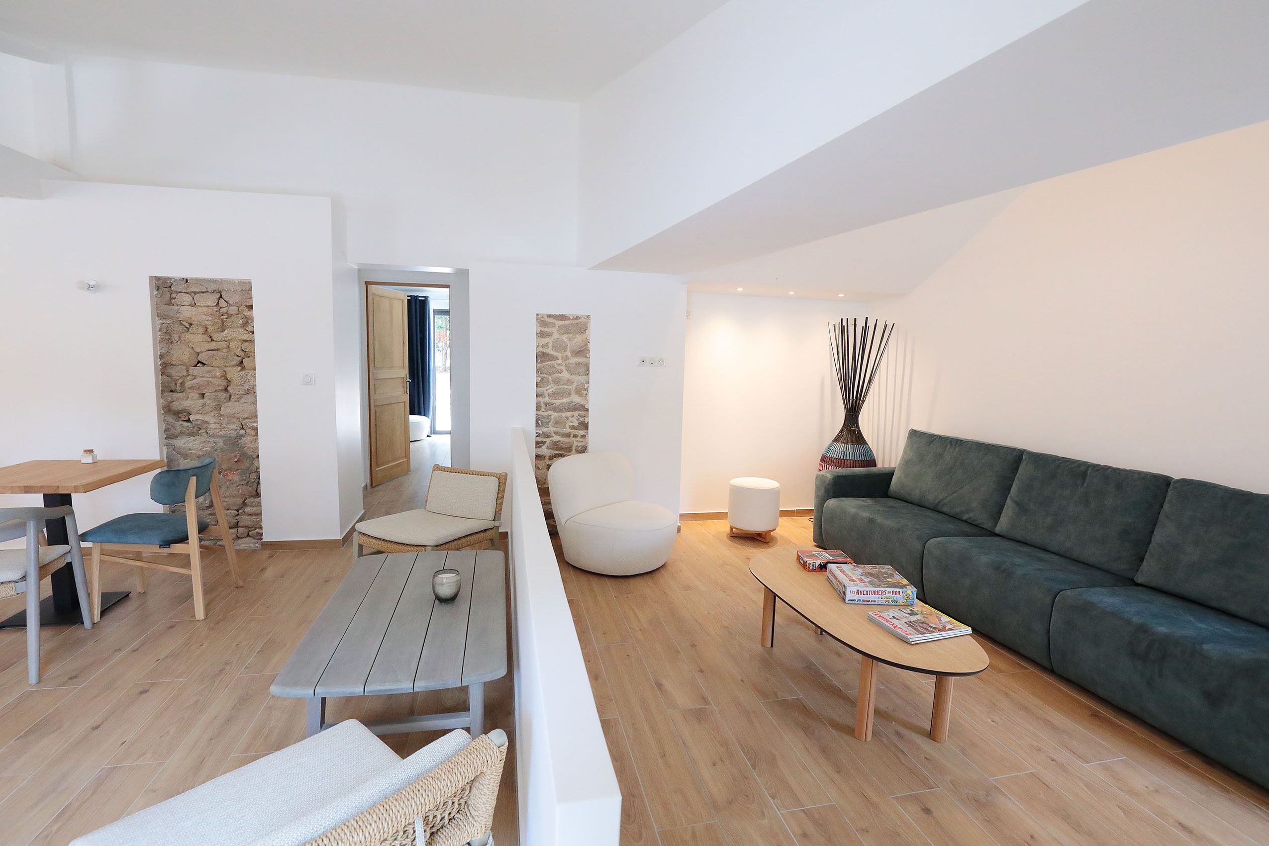 A modern living room with a wooden floor, a green sofa, a white armchair, a round white chair, a wooden coffee table, and a stone wall.