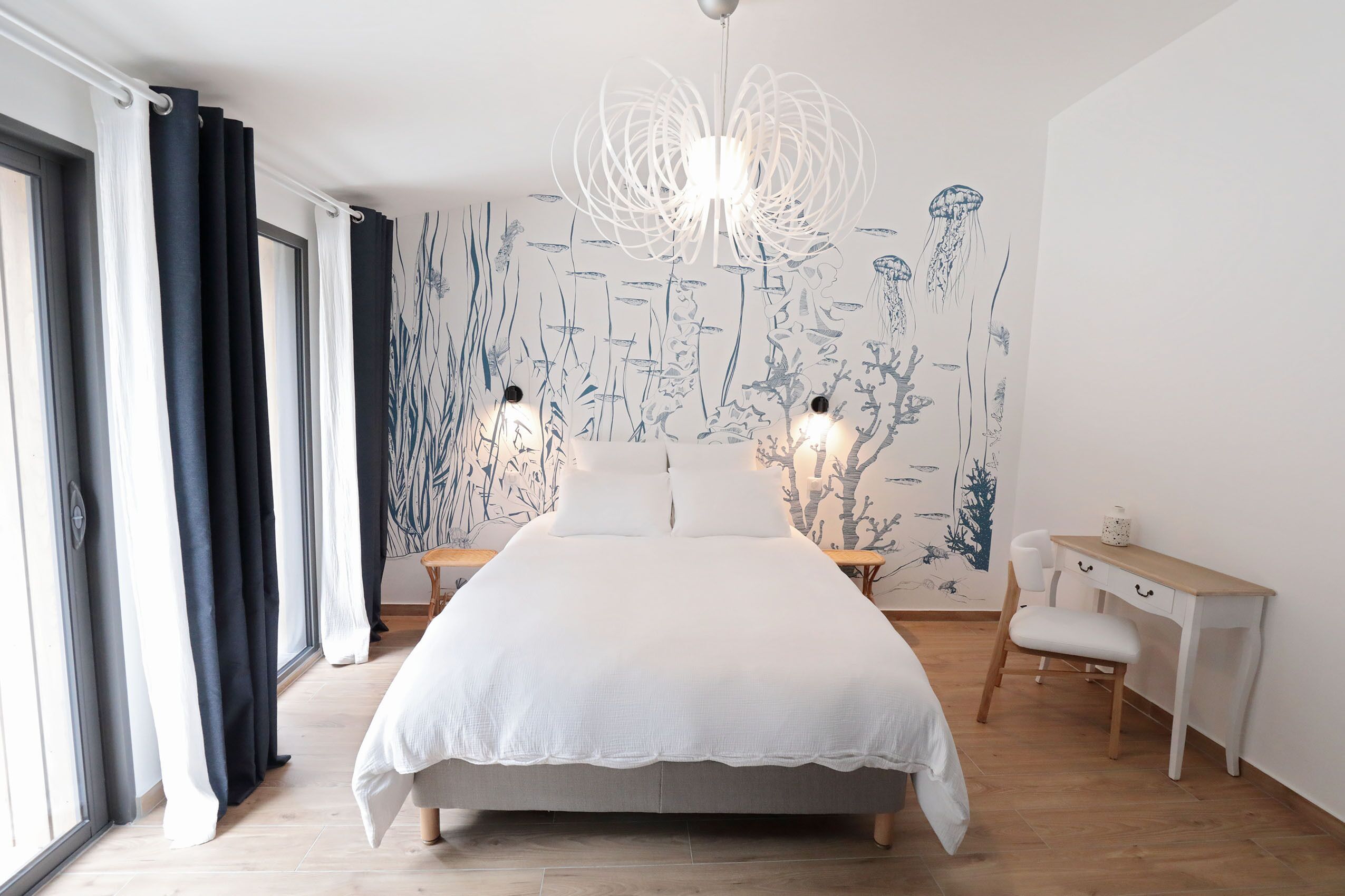 Une chambre avec un grand lit, un bureau et une chaise. Il y a une fenêtre avec des rideaux, un mur orné d’une fresque végétale et un luminaire suspendu.