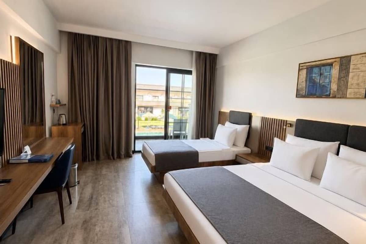 A hotel room with two beds, a desk, a chair, a large window with curtains, and a painting on the wall.
