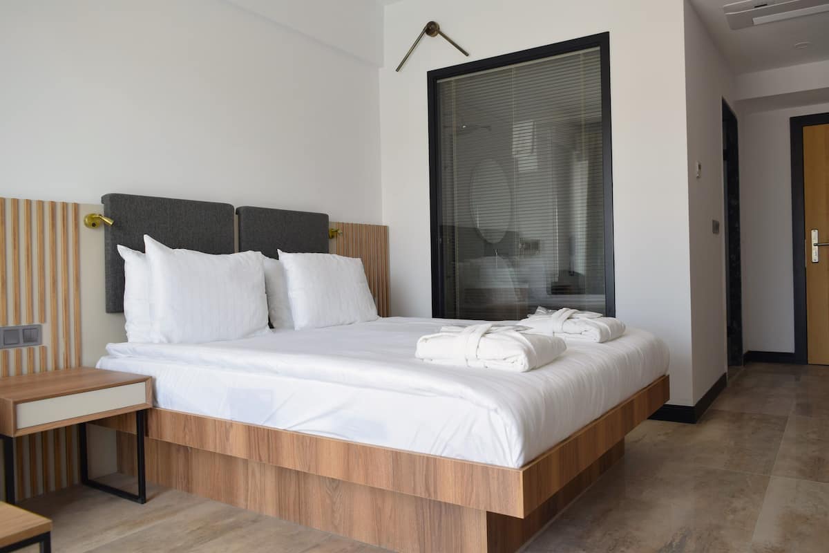 Une chambre d’hôtel moderne avec un lit en bois, du linge de lit blanc, une table de chevet et une grande fenêtre avec des stores.